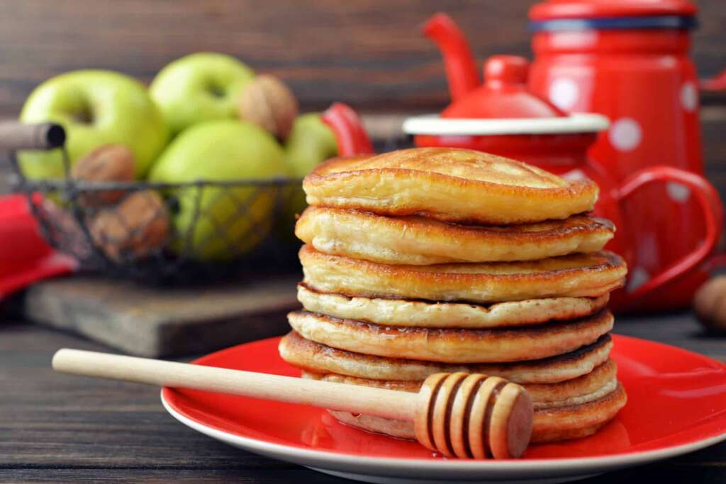Stack of pancakes on plate with vintage teapots and fresh apples on wooden background